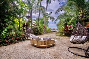 A circular wicker sofa with cushions, two hanging wicker chairs, and a garden path with tropical plants and a view of the sea.