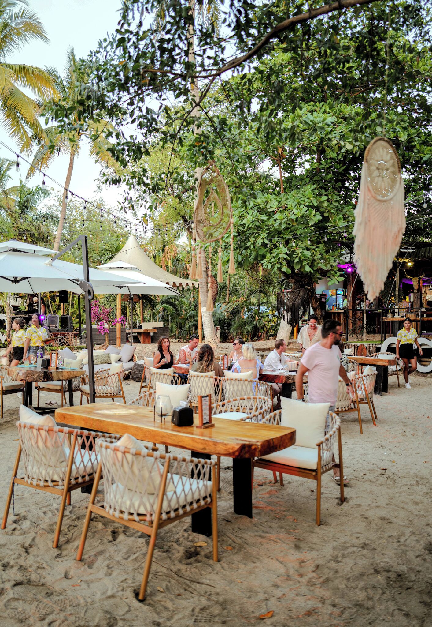 Outdoor seating with tables and chairs, surrounded by trees and umbrellas.