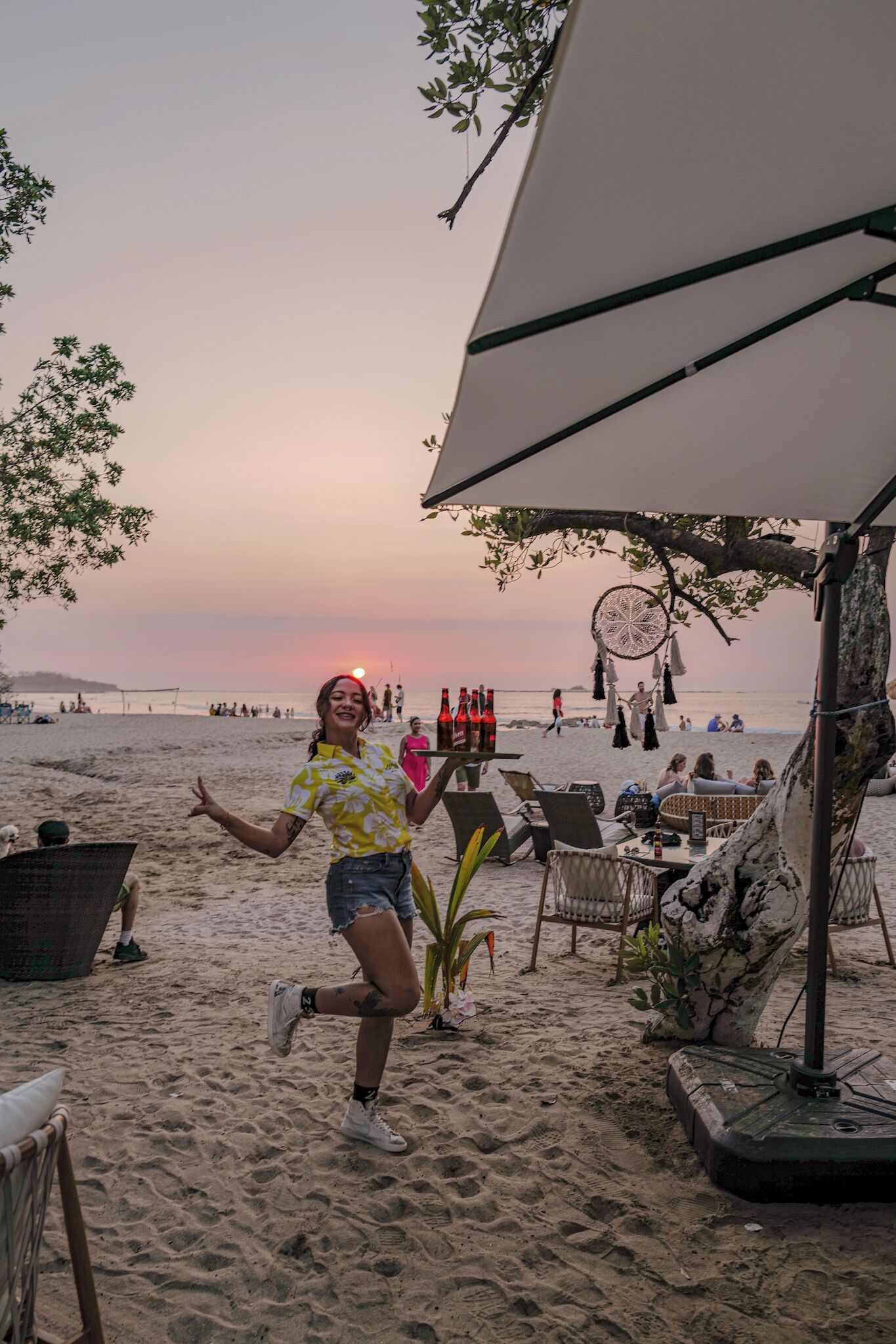 Un cadre en bord de mer, avec une personne debout près d’une table sur laquelle se trouvent des bouteilles, entourée de chaises de plage et d’un parasol.