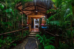 L’entrée d’un chalet tropical, avec un toit de chaume, des rampes en bois et un chemin de pierre menant à une pièce comprenant un lit et une petite table.