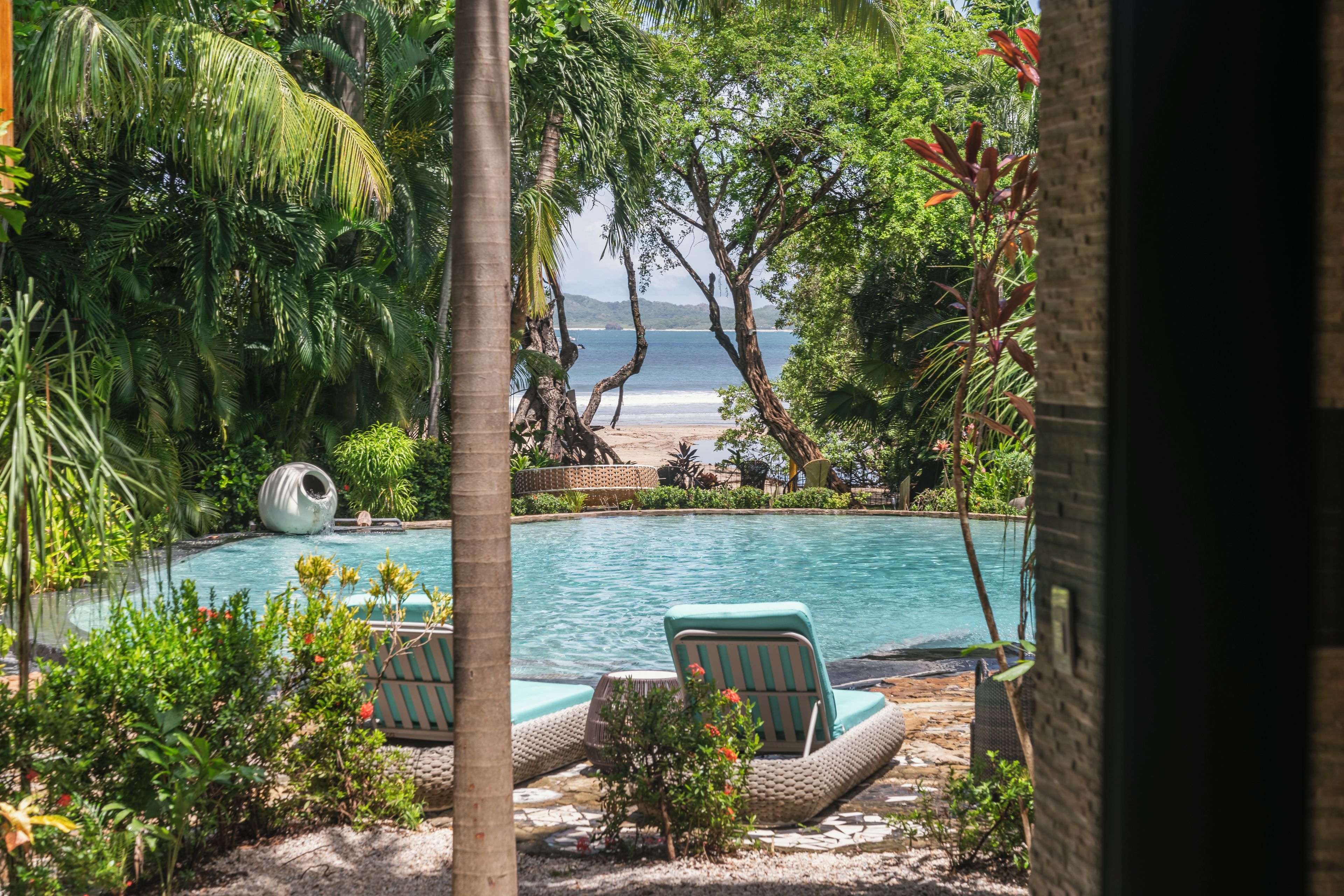 A pool with lounge chairs, surrounded by tropical vegetation and a view of the beach.
