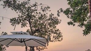 Een strandsetting met rieten loungezetels en parasols, een boom die schaduw biedt, en een zonsondergang op de achtergrond.