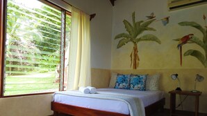 A room with a bed, a window with blinds, a wall mural, and a bedside table with lamps. - Diquis Del Sur (Ciudad Cortés)