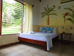 A room with a bed, a window with blinds, a wall mural, and a bedside table with lamps. - Diquis Del Sur (Ciudad Cortés)