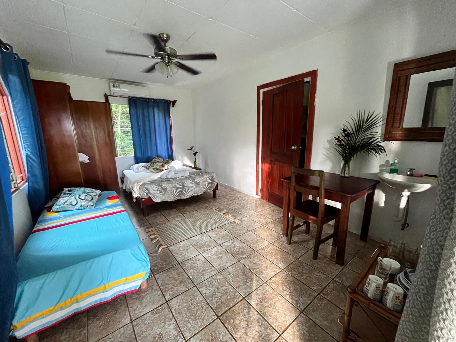 A room with a bed, a desk, a chair, a ceiling fan, a door, and a window with curtains.