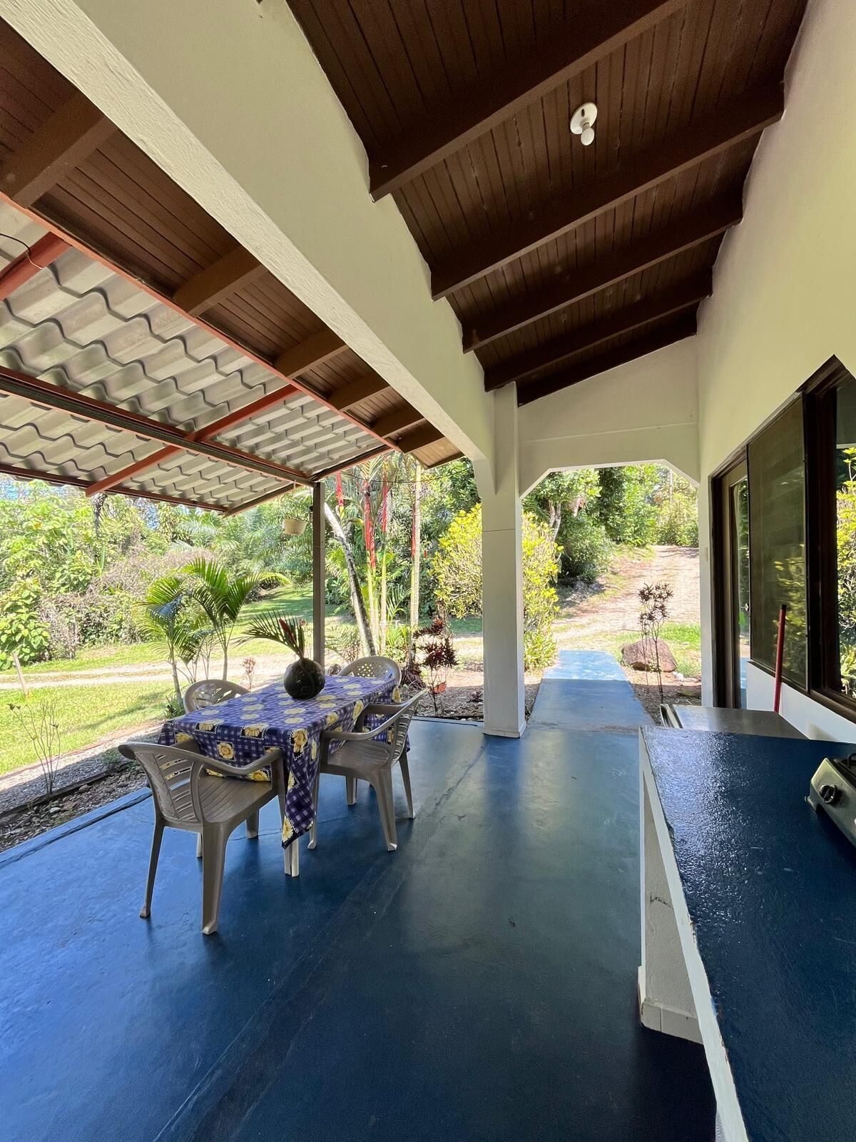 An outdoor covered patio with a table and chairs, a view of a garden, and a blue floor.