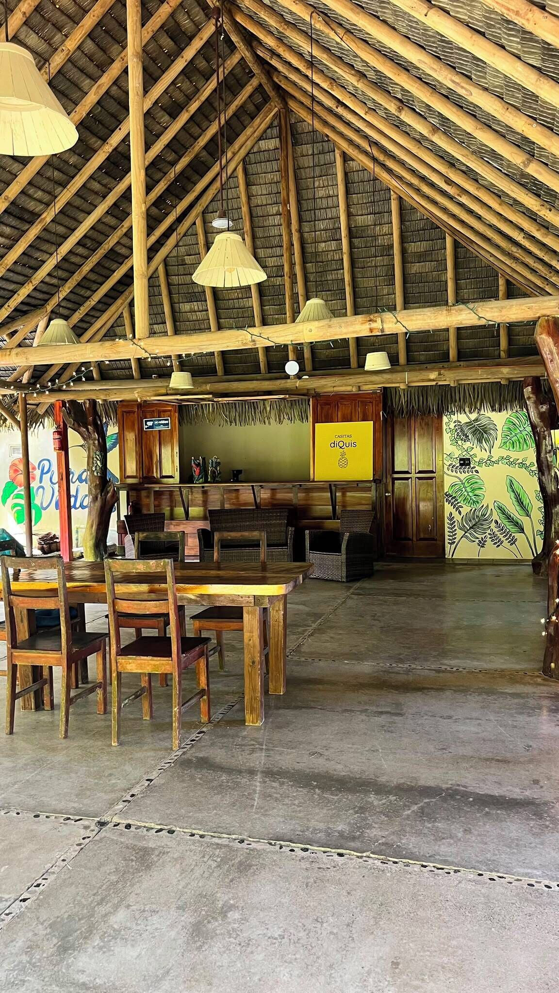 Un espacio interior con techo alto, vigas de madera y tejado de paja. Hay un comedor con muebles de madera y lámparas colgantes.