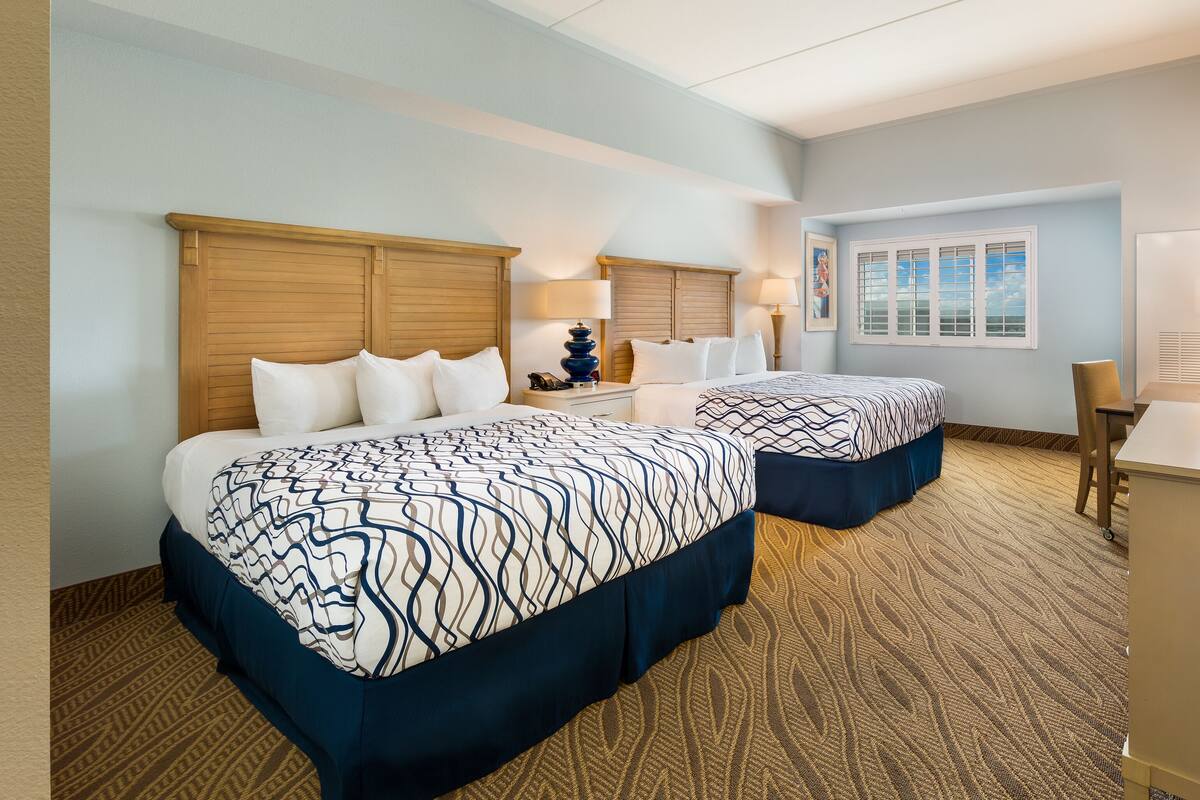 A hotel room with two beds, a desk, a chair, a window, and a wall-mounted TV.