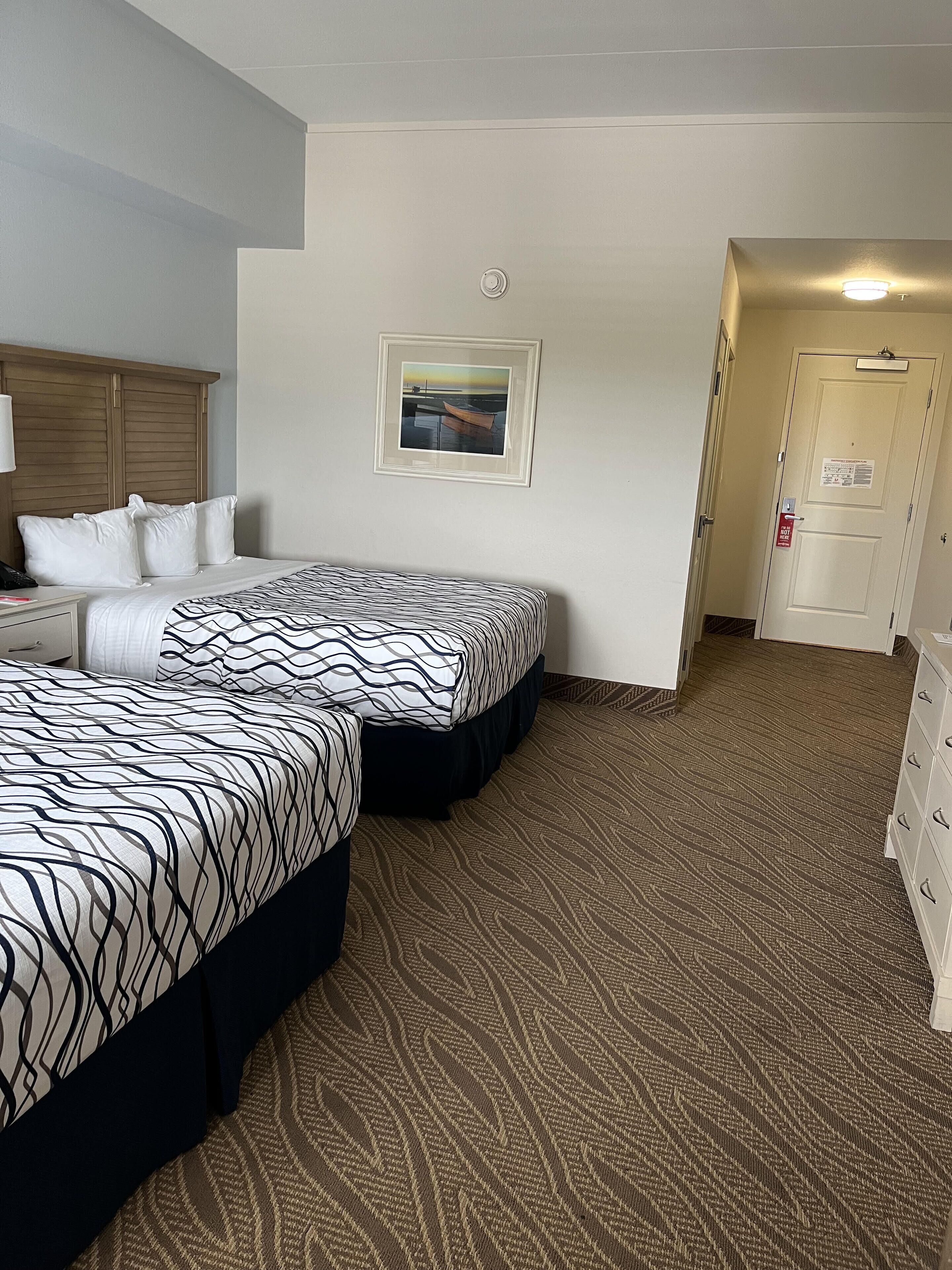 A hotel room with two beds, a wooden headboard, a framed picture on the wall, and a door with a fire safety sign.