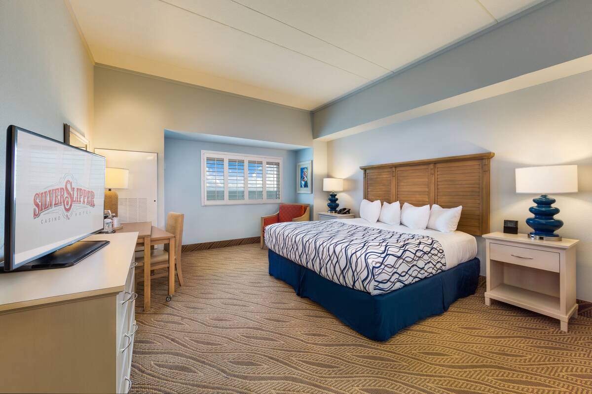 A hotel room with a large bed, a desk with a television, a chair, a nightstand with a lamp, and a window with blinds.