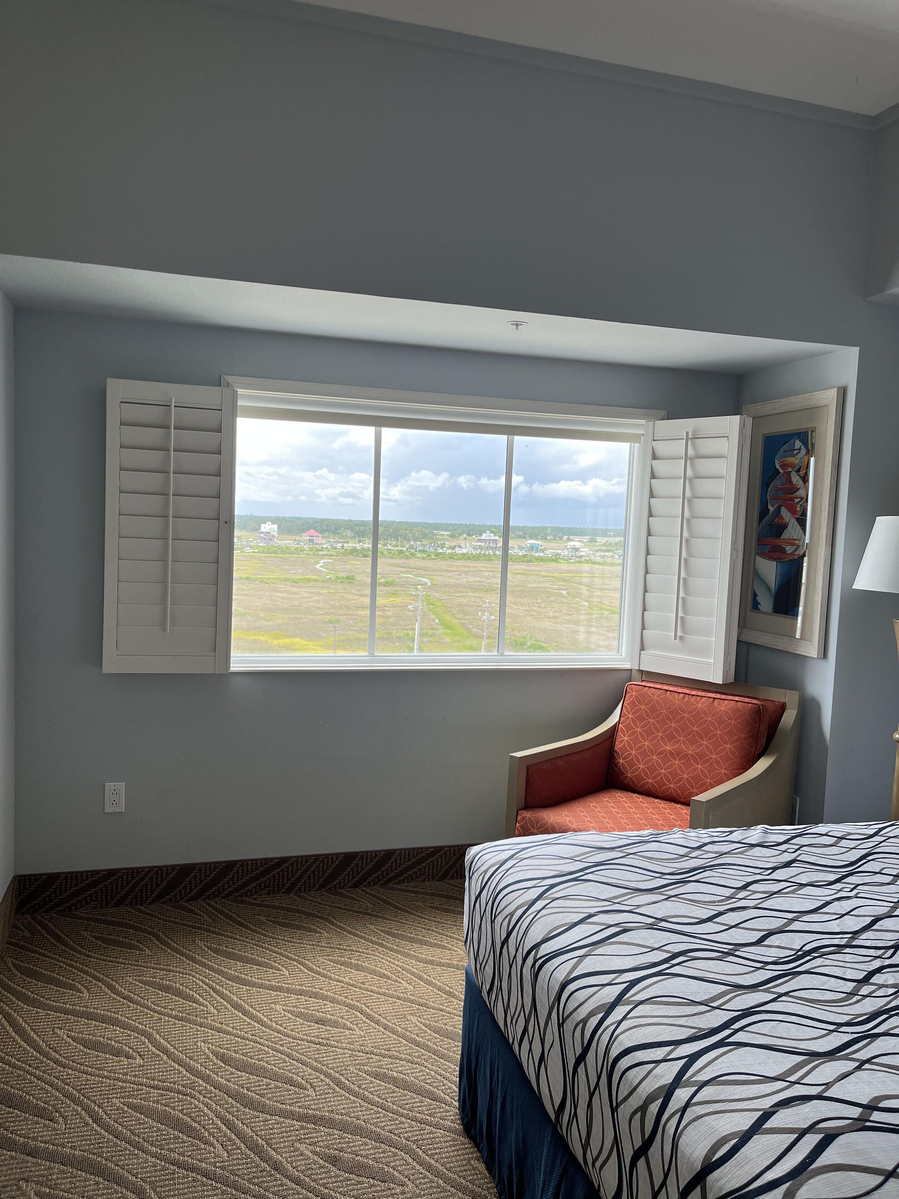 A hotel room with a large window, a bed with patterned bedding, a chair with a geometric cushion, and a painting on the wall.