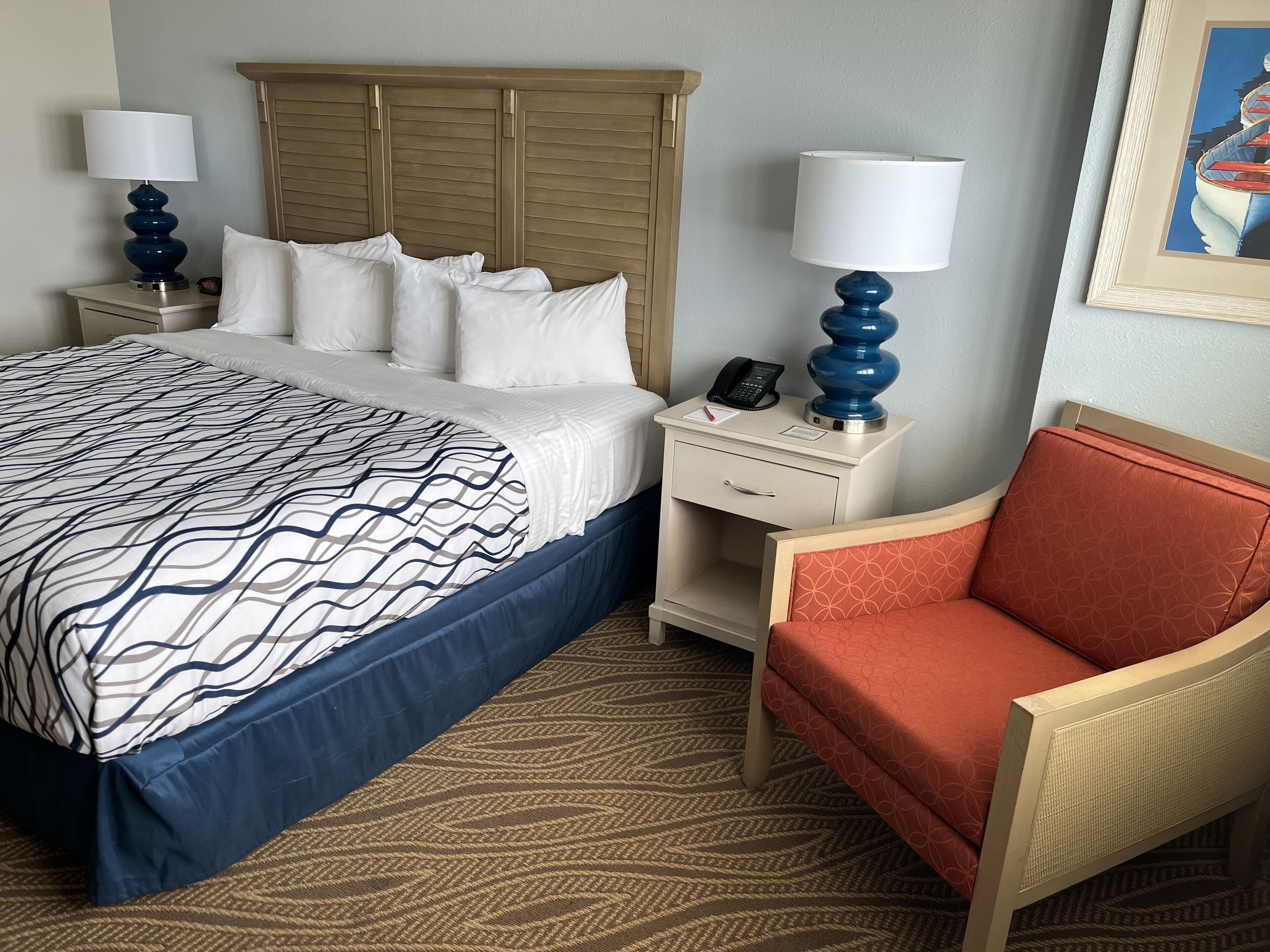 Habitación de hotel con una cama grande, una mesita de noche con lámpara, una silla y un cuadro en la pared.
