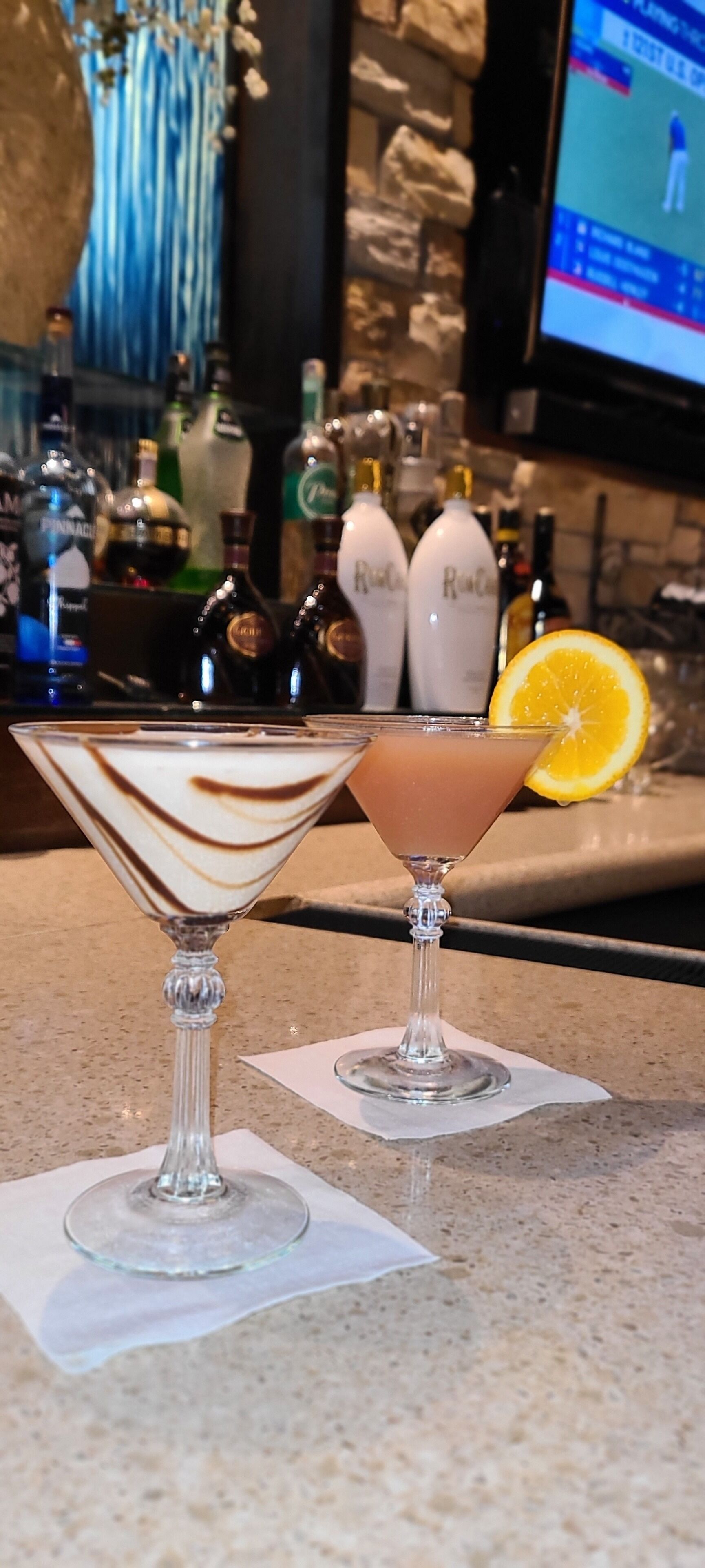 Due bicchieri di martini su un bancone del bar, con una parete in pietra e un televisore sullo sfondo.