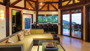 Uma sala de estar ampla com um sofá grande, uma mesa de centro de vidro e vista para o exterior através de grandes portas deslizantes.