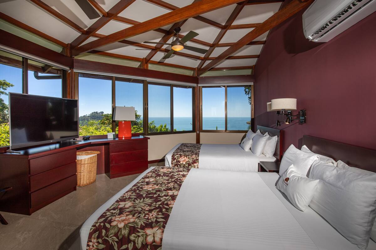 Habitación de hotel con un televisor grande, un escritorio, una cama con ropa de cama floral y vistas al mar.