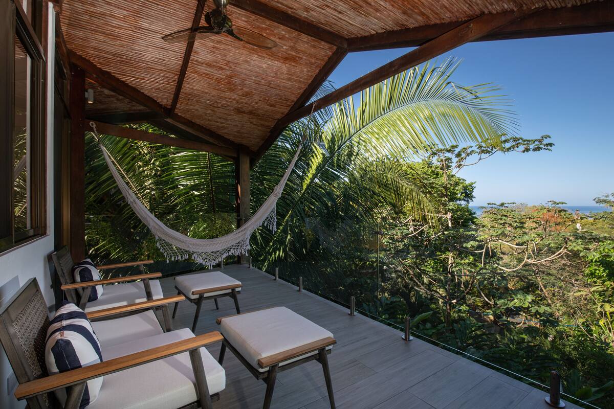 Un espacio de descanso al aire libre con mobiliario de madera y una hamaca, con vistas a un paisaje exuberante y el océano.