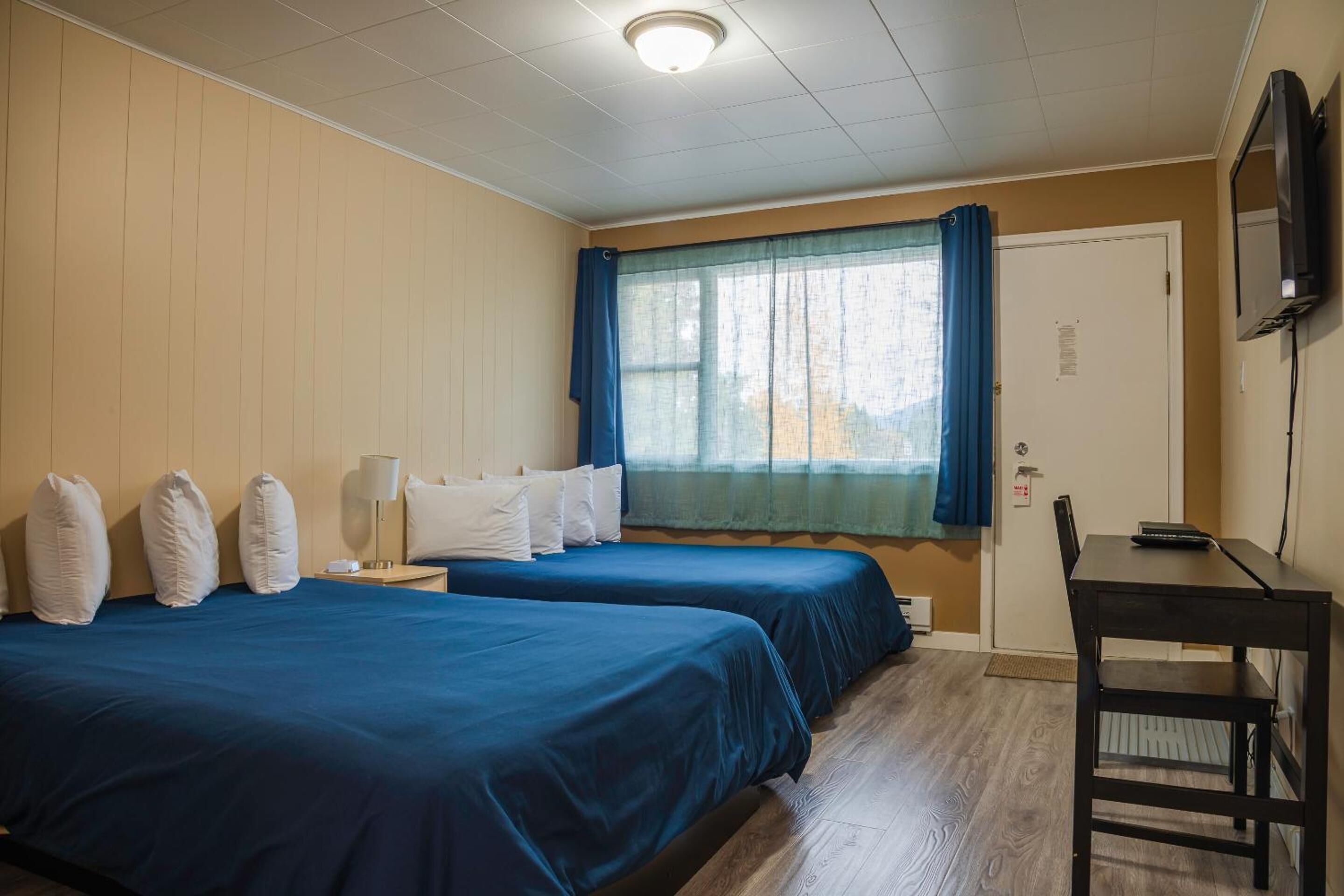 Ein Hotelzimmer mit zwei Betten, einem Schreibtisch, einem Stuhl, einem Fernseher und einem Fenster mit Vorhängen.