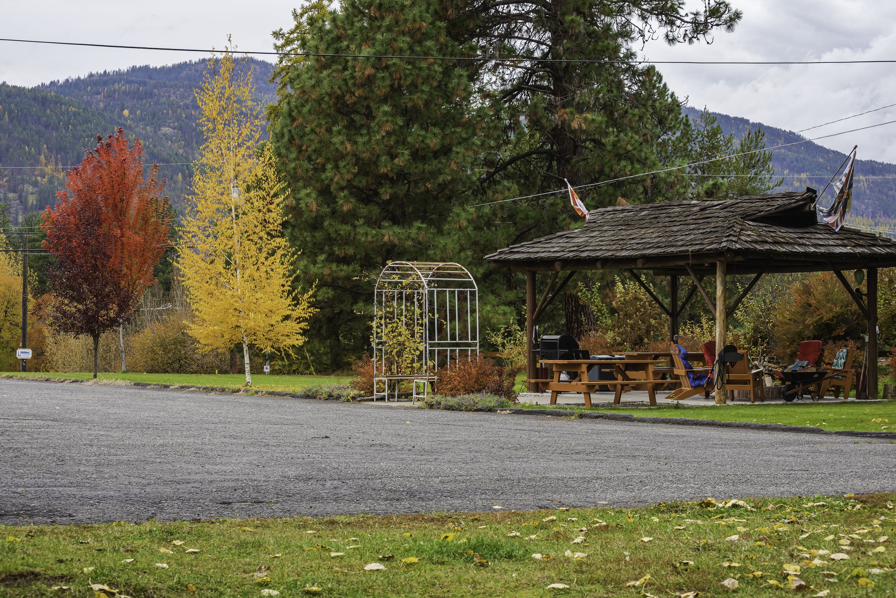 Um gazebo com bancos e um pequeno jardim, cercado por árvores e uma montanha ao fundo.