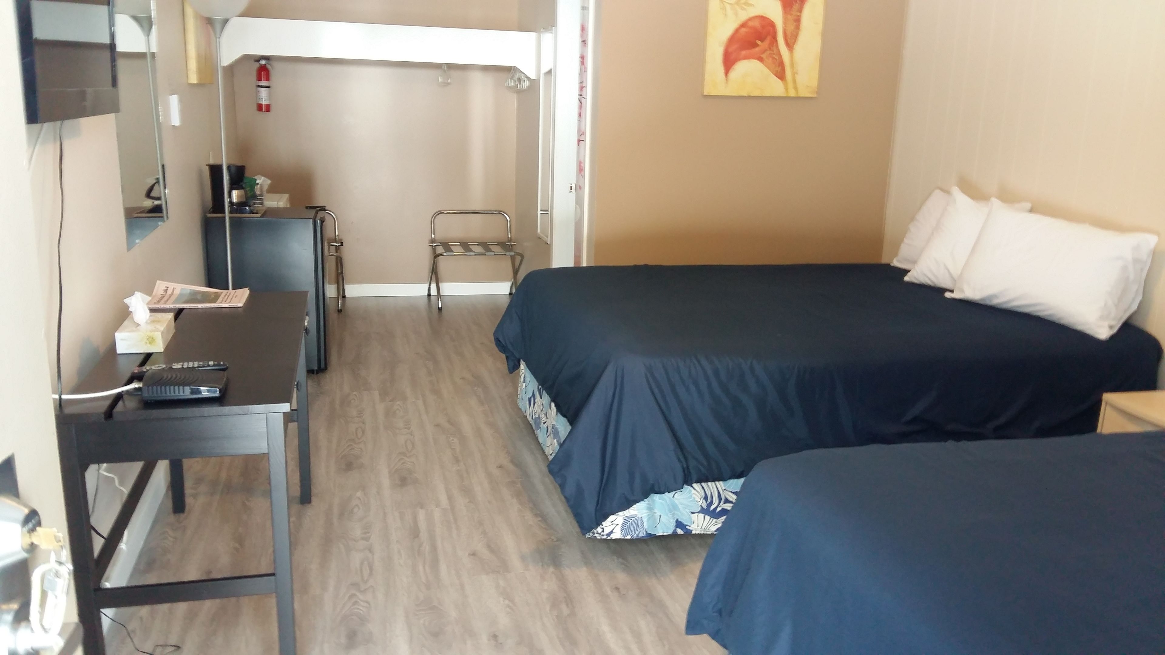 A hotel room with two beds, a desk with a chair, a mirror, a fire extinguisher, and a painting on the wall.
