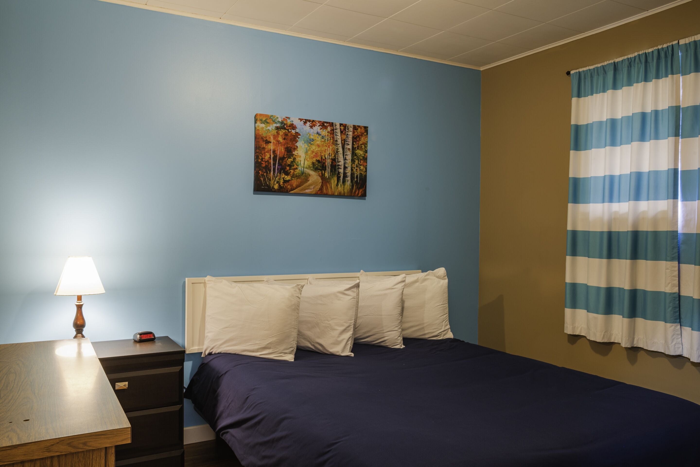 Un dormitorio con cama, mesita de noche, lámpara, un cuadro y una ventana con cortinas.