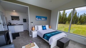 Une chambre moderne avec un grand lit, une vue sur une forêt par la fenêtre et une kitchenette dans la pièce adjacente.