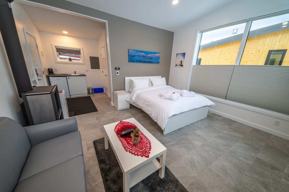 A modern hotel room with a bed, a small desk, a chair, a kitchenette, and a large window with blinds.