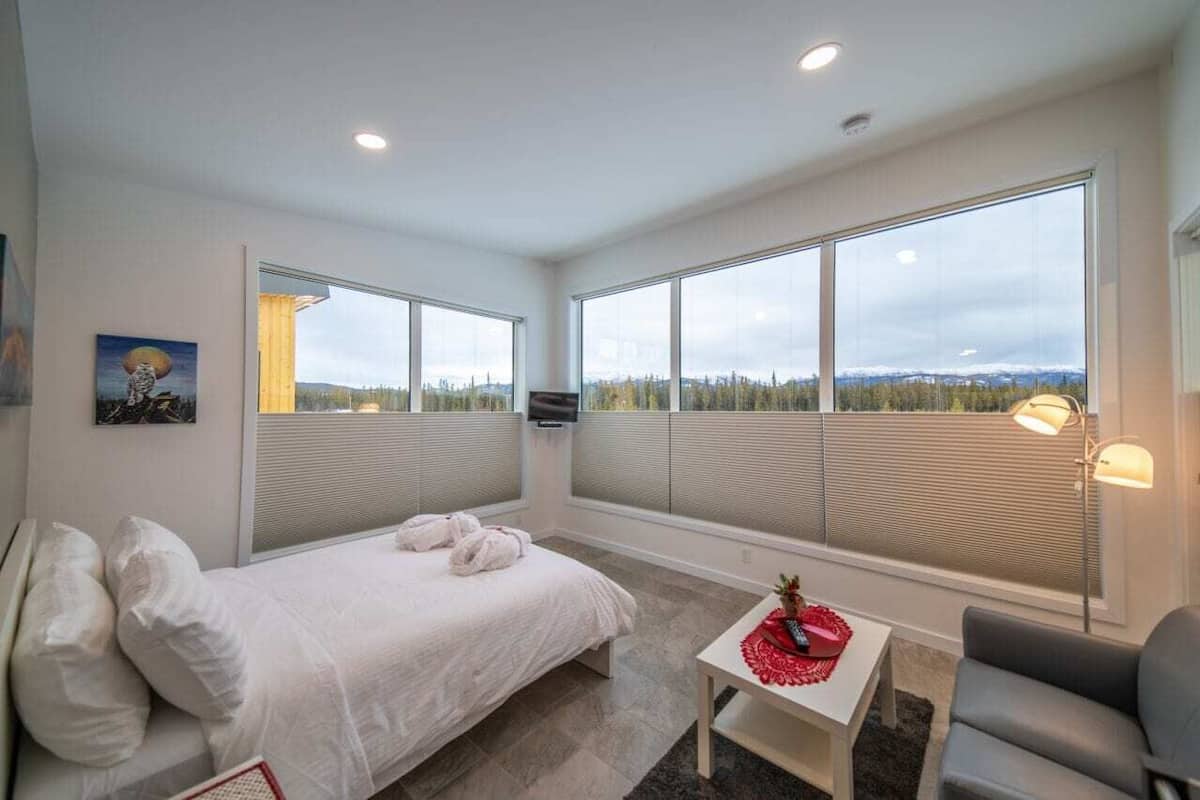 A modern hotel room with a large bed, a small table with a red decorative item, a gray armchair, and large windows offering a view of the landscape.