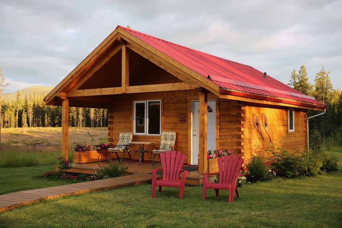 A log cabin with a red roof, two Adirondack chairs, and a small porch.