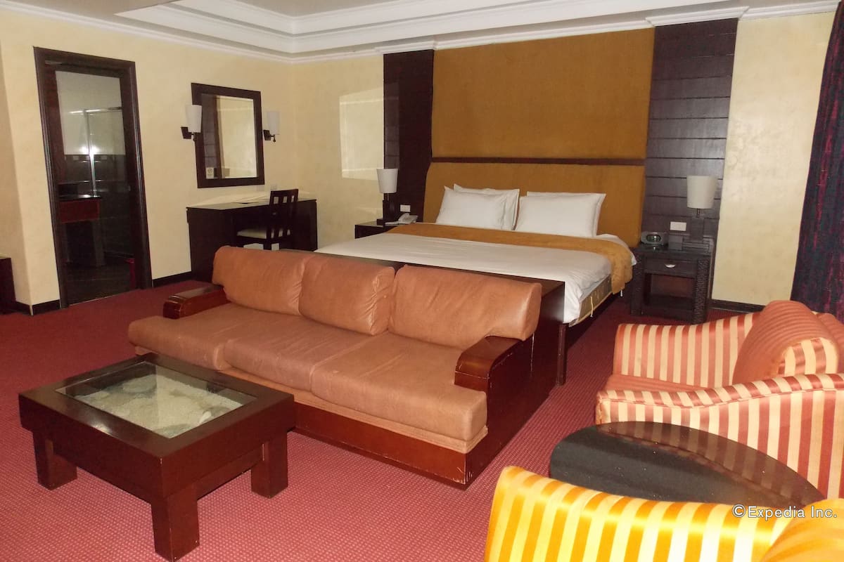 Habitación de hotel con cama, sofá de cuero, sillón a rayas, mesa de centro de vidrio, escritorio y espejo.