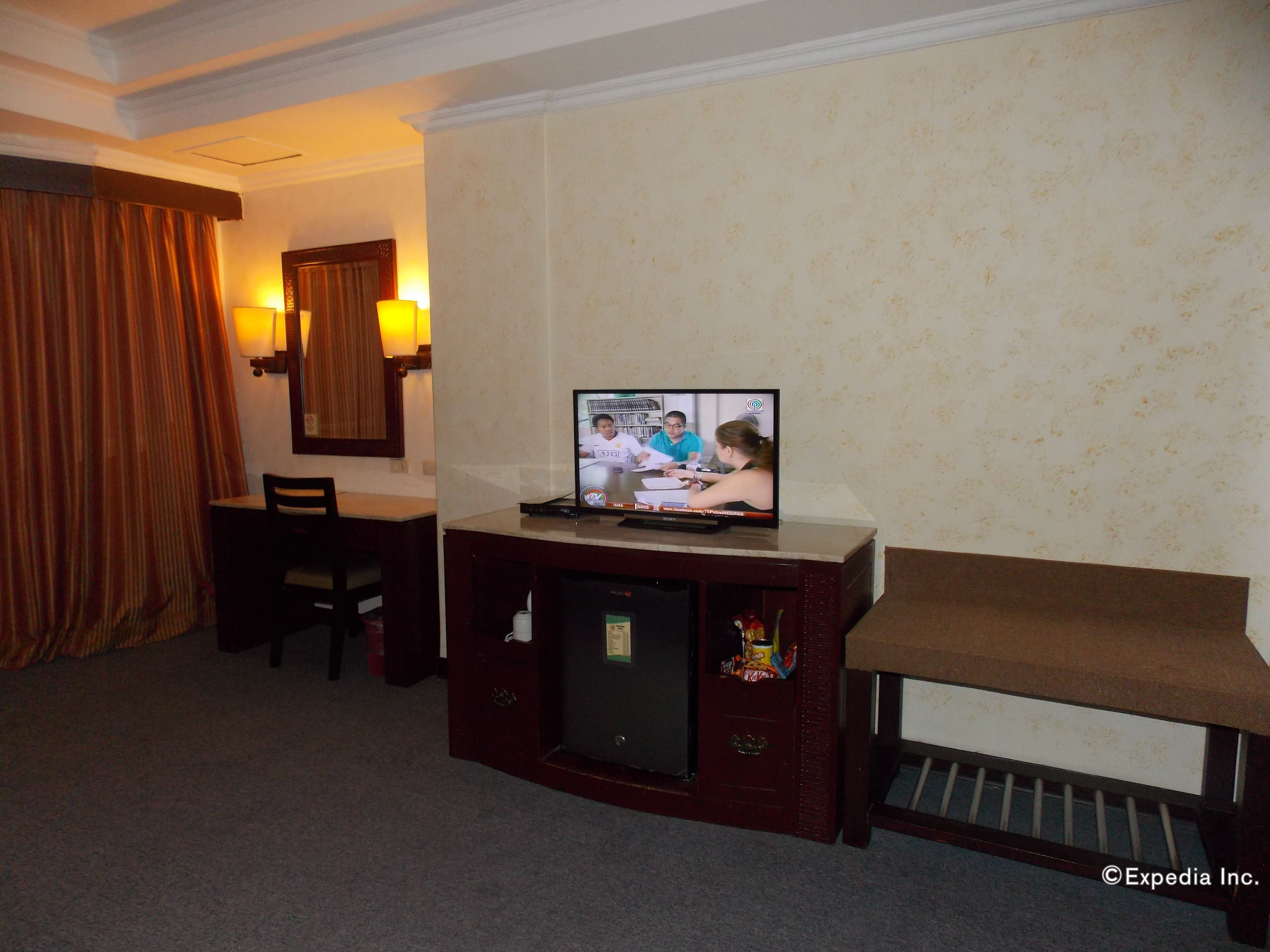 A hotel room with a television on a wooden stand, a desk, a chair, and a small refrigerator.