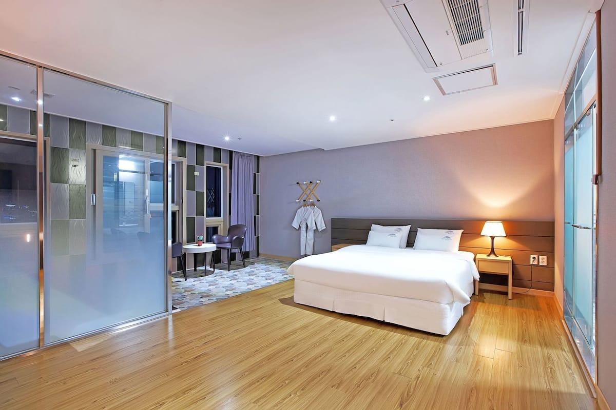 A modern hotel room with a large bed, a desk, a chair, a small table, and a glass partition.