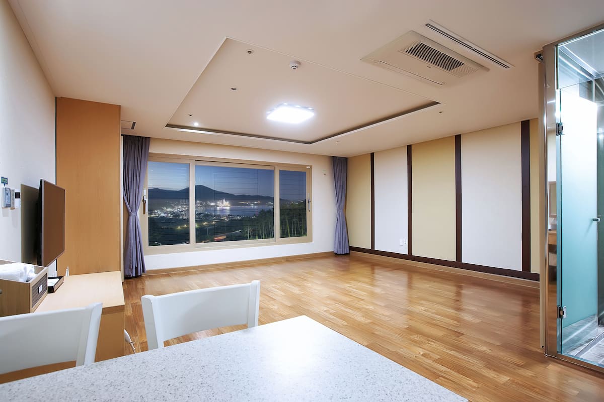 A hotel room with a large window, wooden flooring, a white table, and a TV.