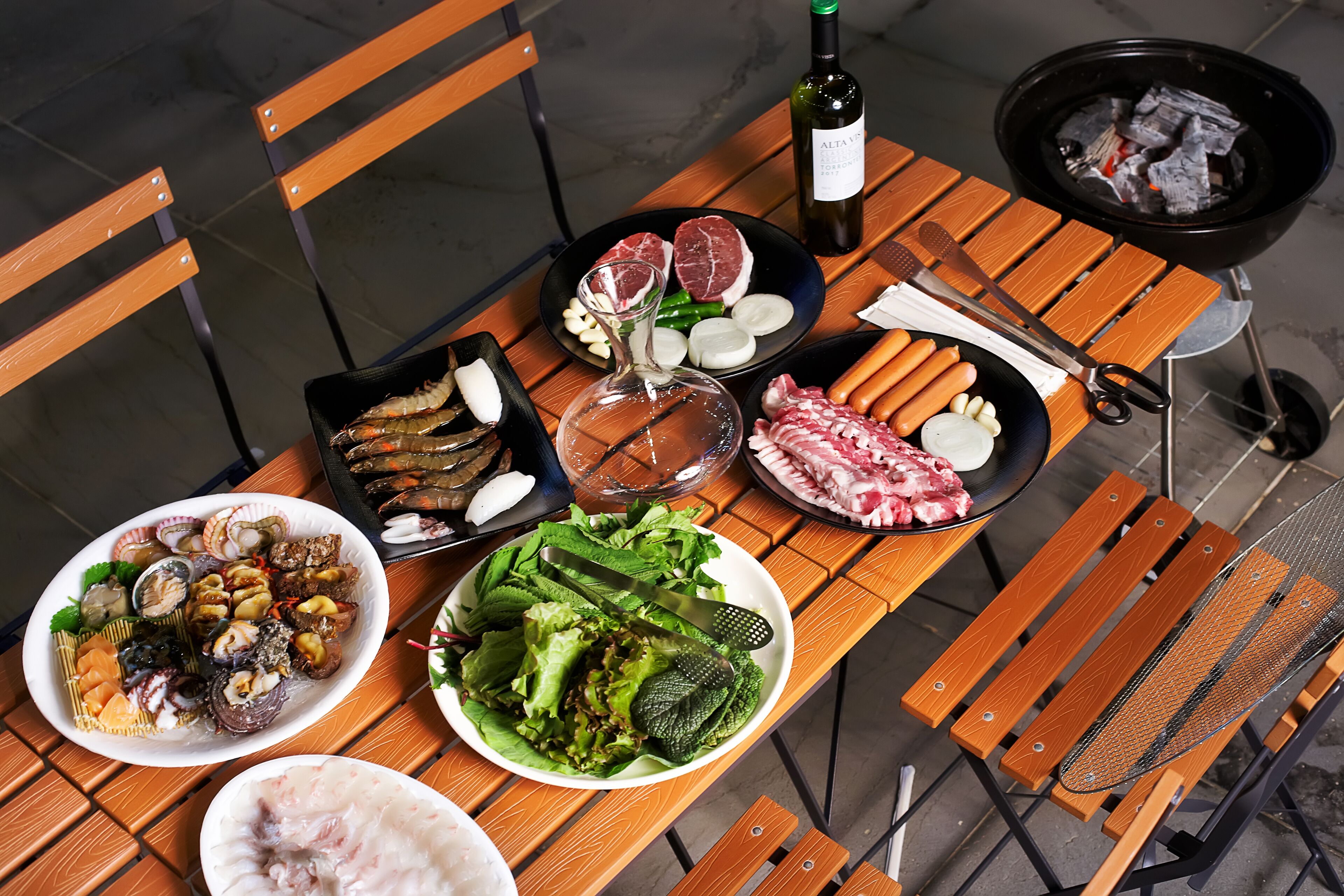 A wooden table with various dishes, including seafood, meat, and vegetables, along with a bottle of wine and grilling utensils.