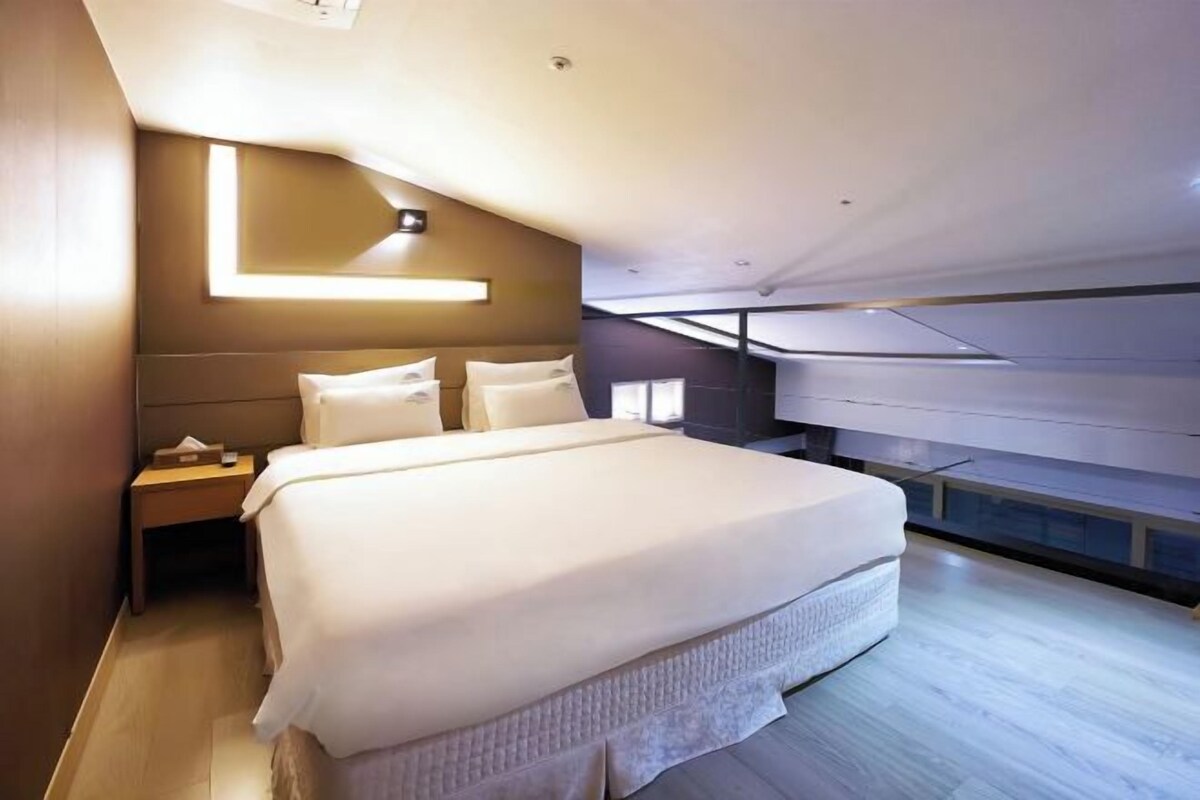 A modern hotel room with a large bed, a nightstand, a wall-mounted light, and a view of the city through a large window.