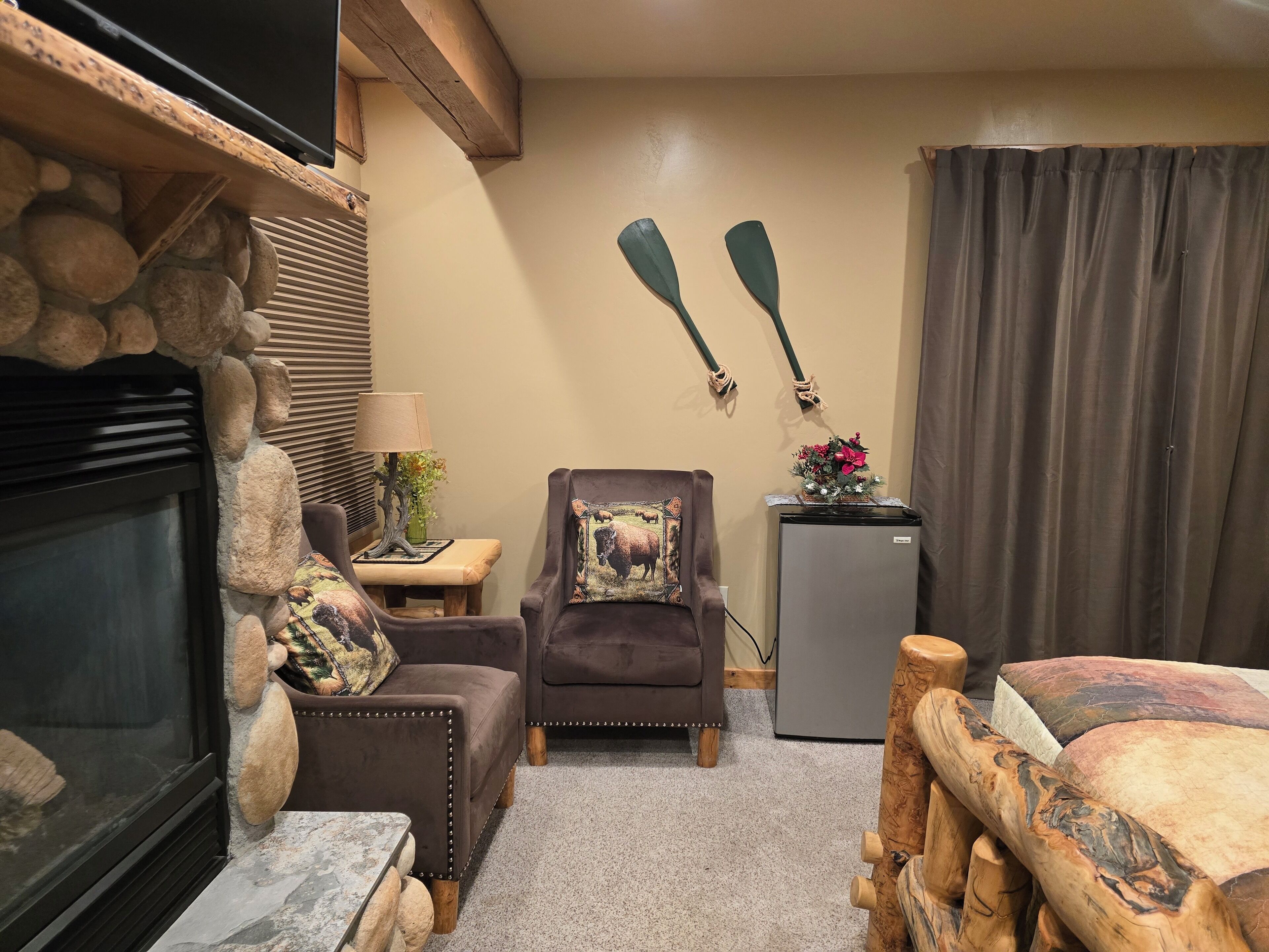 A cozy room with a stone fireplace, a wooden coffee table, a cushioned armchair, a small side table with a lamp, and a refrigerator.