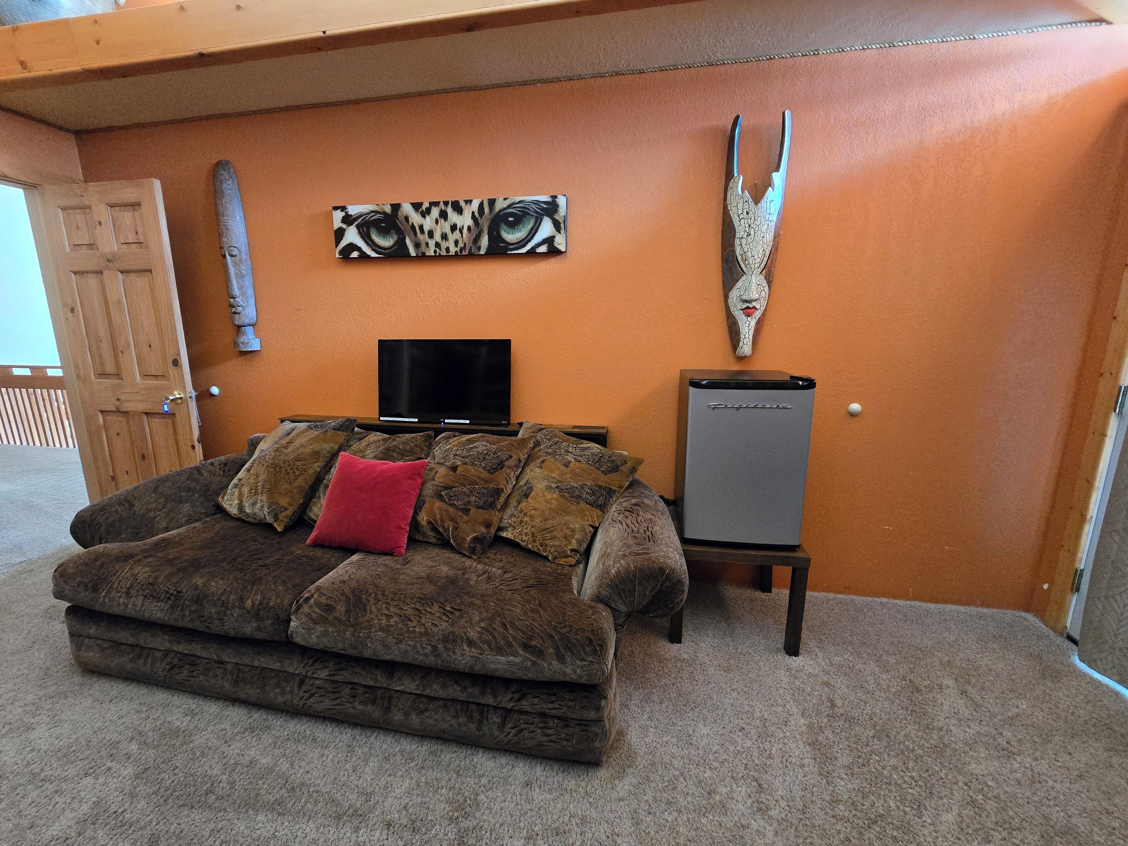 A living room with a brown sofa, a red pillow, a flat-screen TV, and a refrigerator.