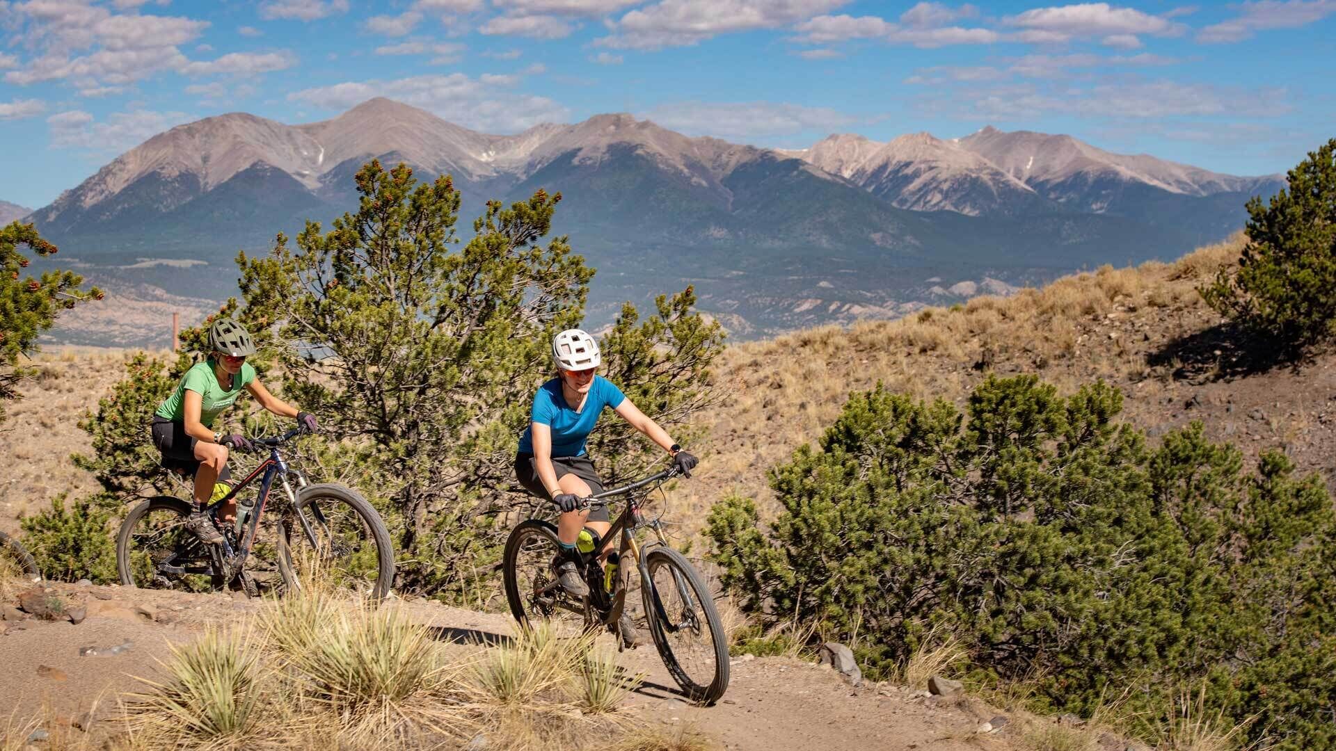 Two cyclists on a mountain trail with a mountainous backdrop.