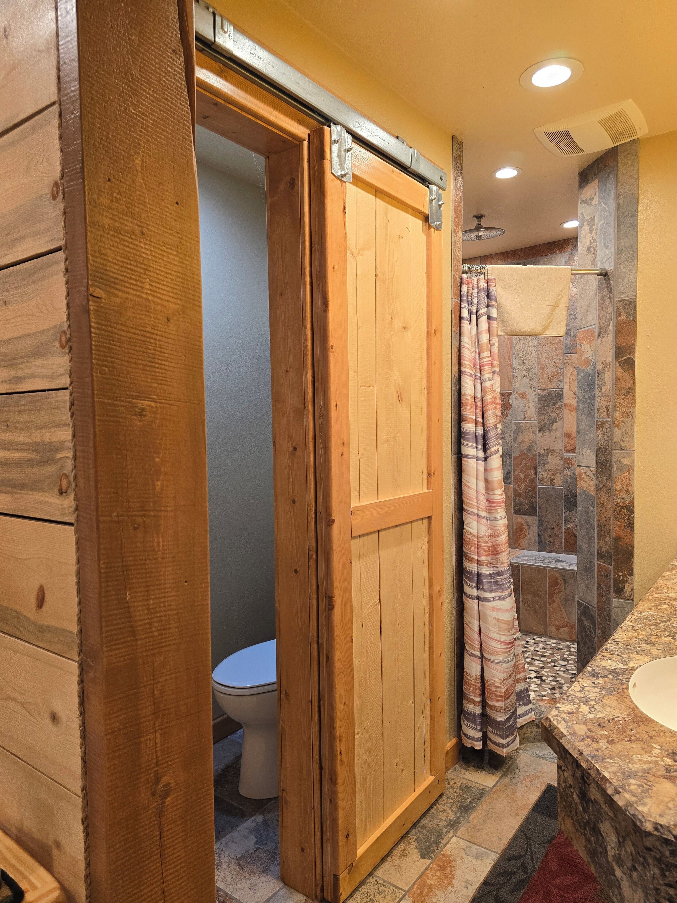 A wooden door leading to a bathroom with a toilet, shower, and a marble sink.