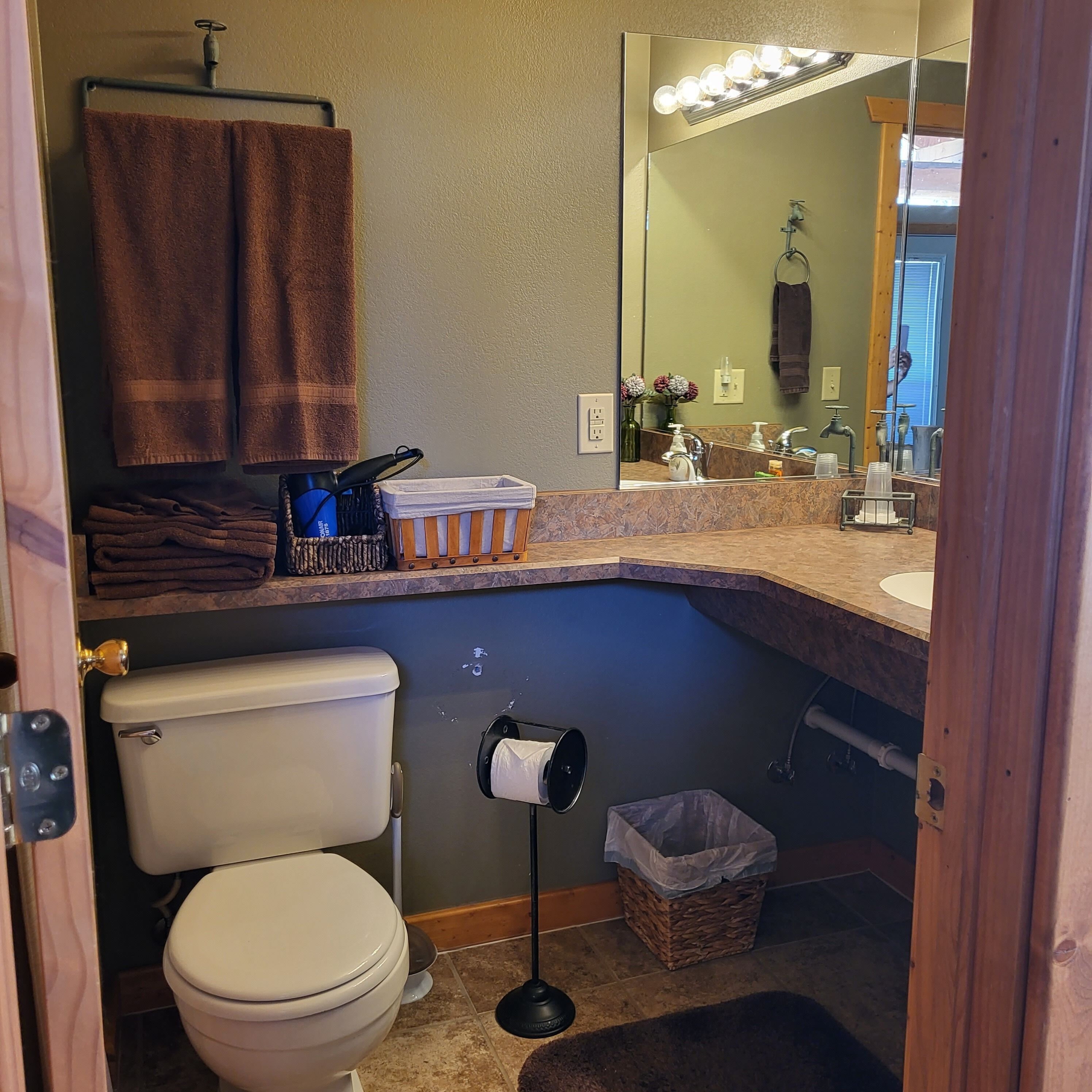 A bathroom with a toilet, sink, and mirror. There are towels hanging on a rack, a trash bin, and a basket.