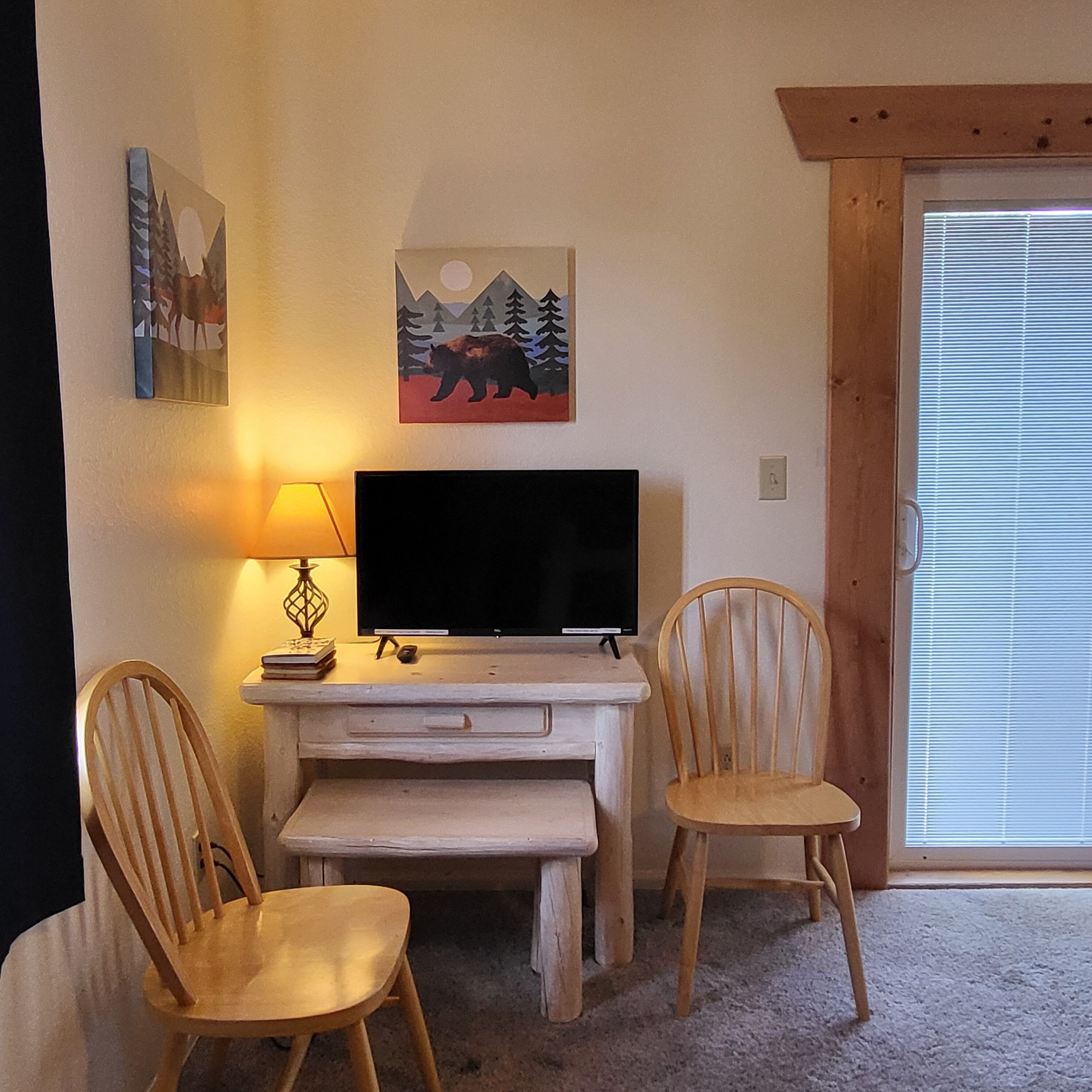 A room with a TV on a wooden desk, two wooden chairs, a lamp, and two paintings on the wall.