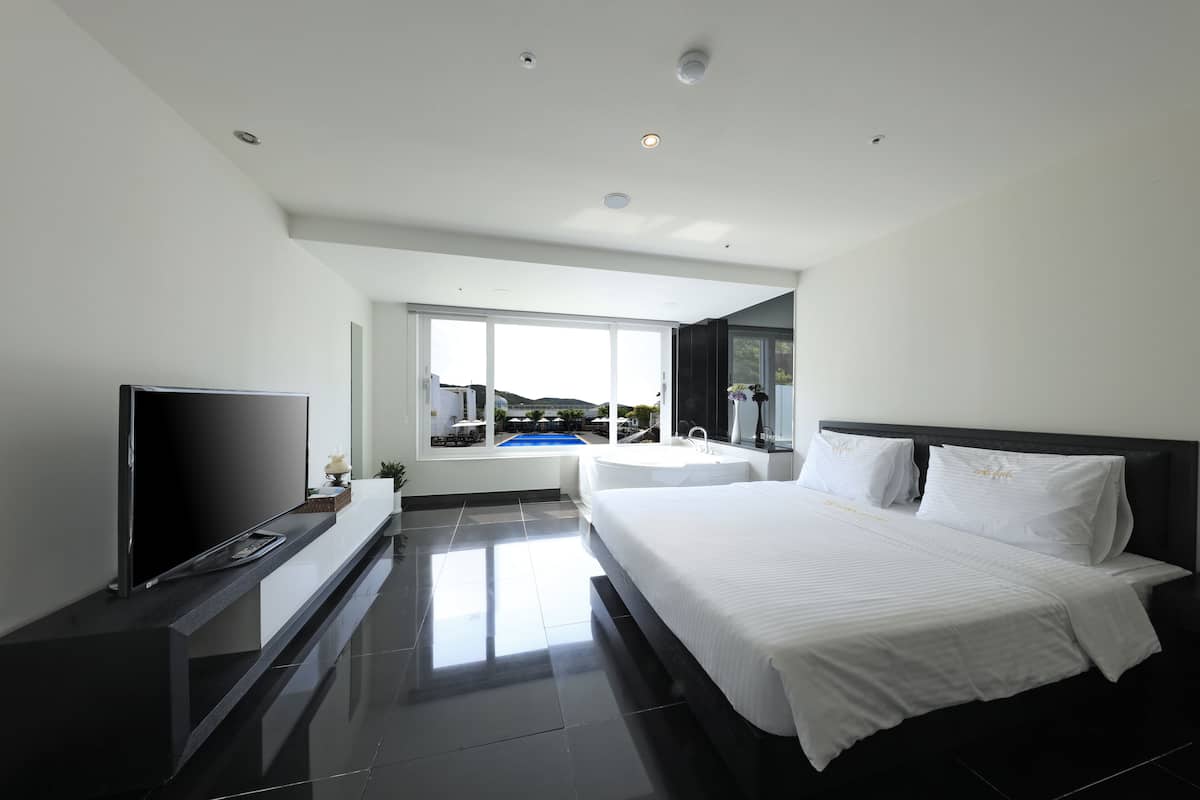 A modern hotel room with a large bed, a flat-screen TV, a bathtub, and a view of a pool.