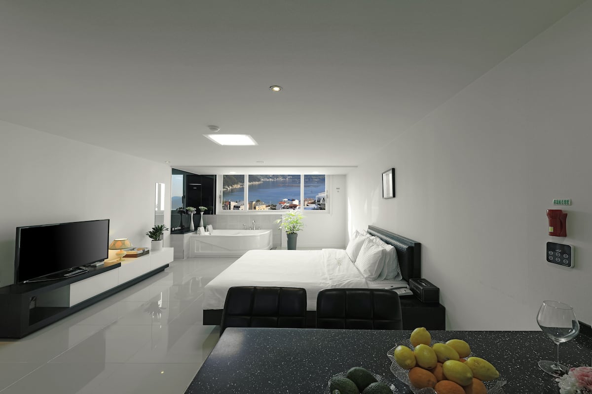 A modern hotel room with a bed, a TV, a dining table with fruit, a view of the sea, and a bathroom with a bathtub.