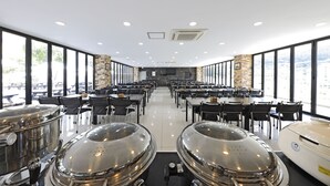 A spacious dining area with large windows, multiple tables, and a kitchen area with stainless steel appliances. - Doya Family Hotel (Geoje)
