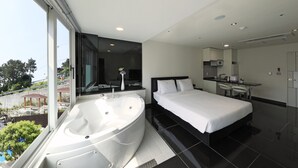A modern hotel room with a large bathtub, a bed, a small kitchenette, and a view of a pool and trees. - Doya Family Hotel (Geoje)