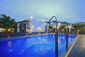 Una piscina con chorros de agua, una terraza de madera y un edificio iluminado al fondo.