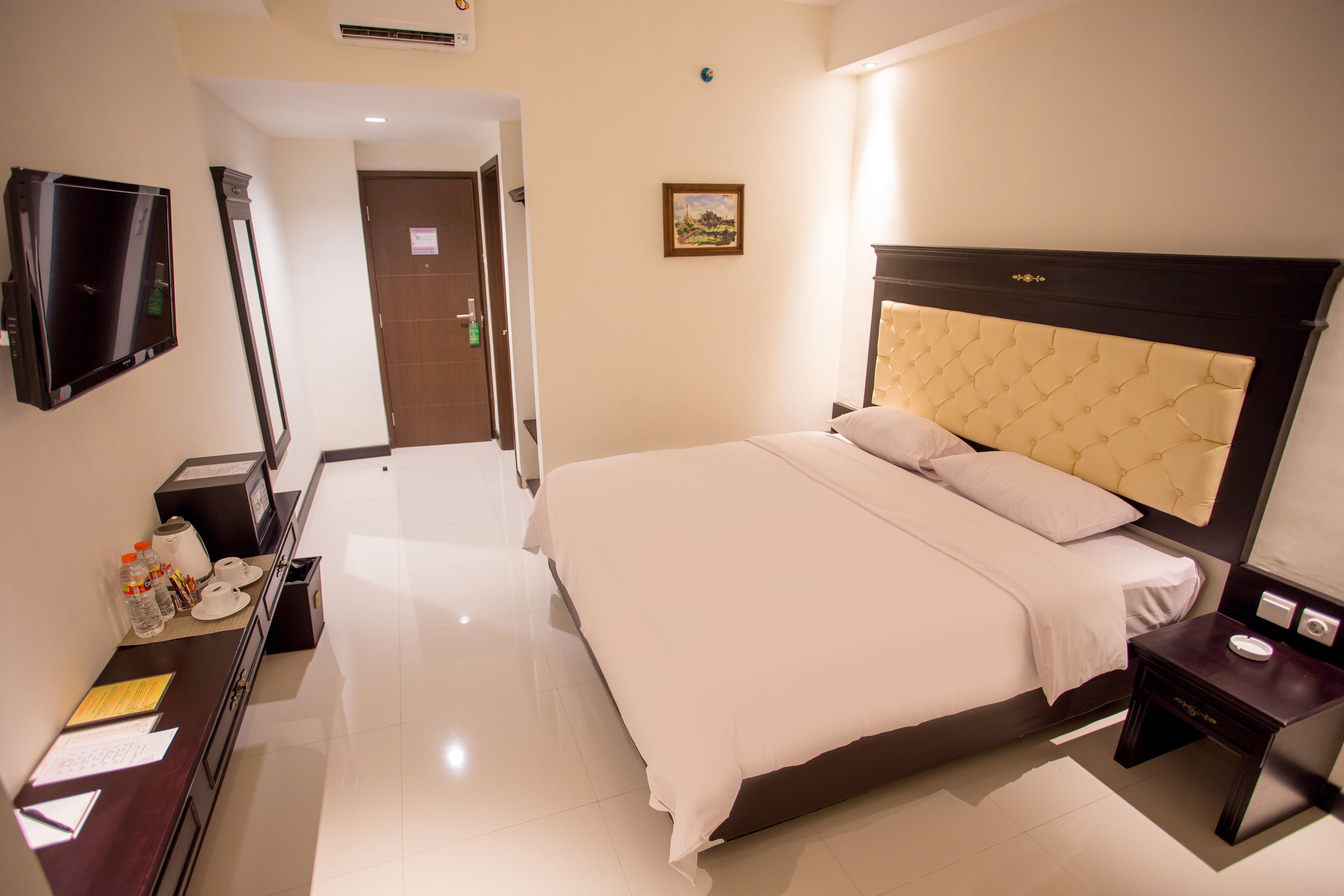 A hotel room with a large bed, a TV mounted on the wall, a bedside table with a lamp, and a small desk with a mirror and some items on it.