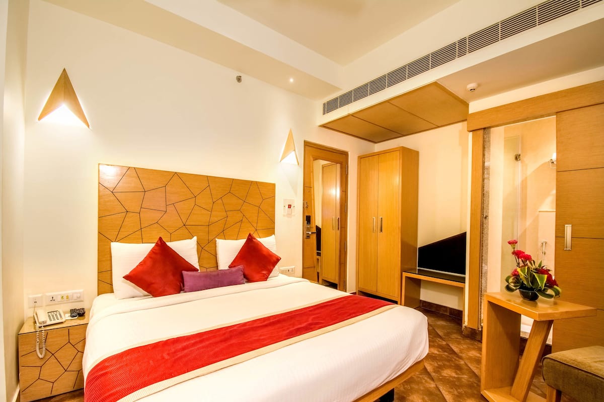 A hotel room with a large bed, wooden headboard, bedside tables, a TV, wardrobe, and a small table with a vase.