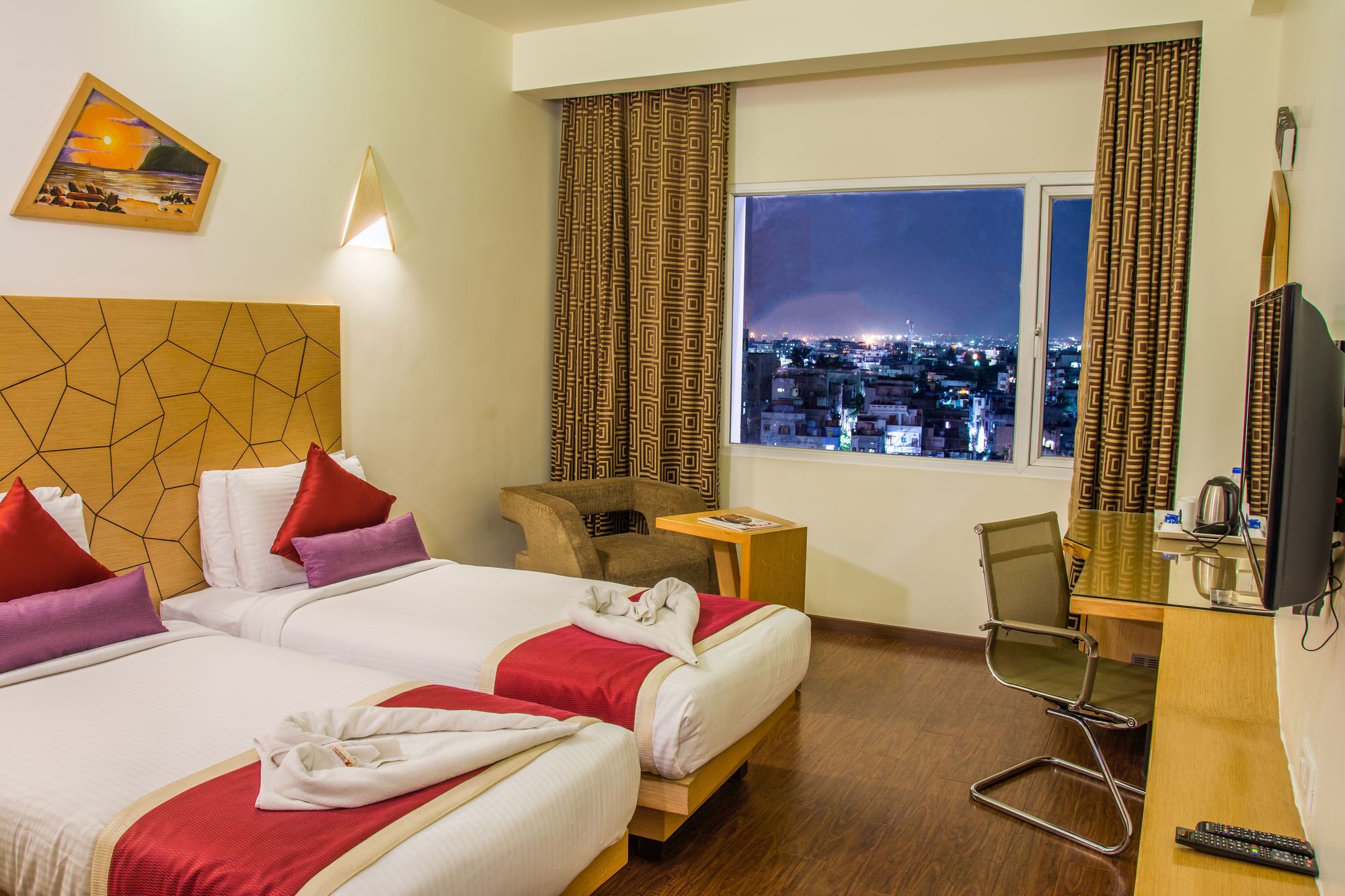 A hotel room with two beds, a desk with a chair, a television, and a city view.