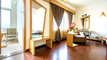 A modern hotel room with a wooden floor, a glass desk, a sofa, a chair, a mirror, and a window with curtains.