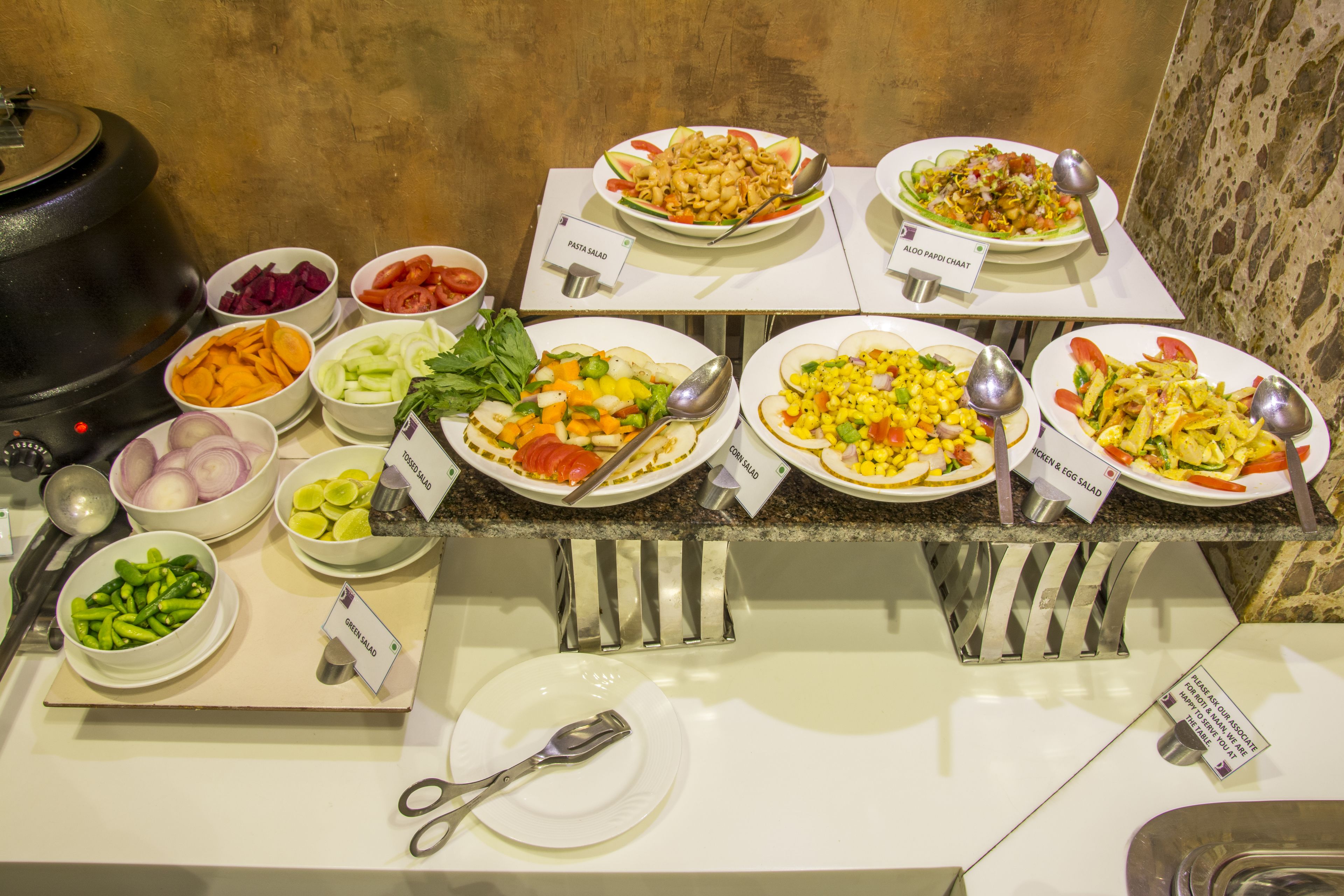 Un buffet proposant divers plats, chacun étant clairement identifié, notamment une salade de pâtes, de la mangue verte et une salade de maïs.
